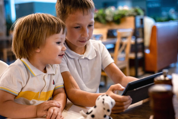 Boy and toddler playing video games on tablet online on internet while waiting orders in restaurant. Children using smartphone. Kids using technology. Screen time addiction. - Foto, immagini