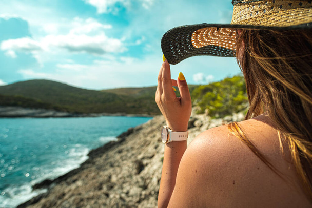 Egy felismerhetetlen barna lány oldalnézete, aki nyáron a szénakalapjába kapaszkodik. Az adriai tenger partjainál állva, az utazás, a nyár és a nyaralás koncepciói - Fotó, kép