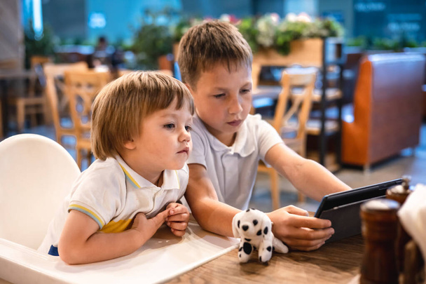 Boy and toddler playing video games on tablet online on internet while waiting orders in restaurant. Children using smartphone. Kids using technology. Screen time addiction. - Фото, зображення