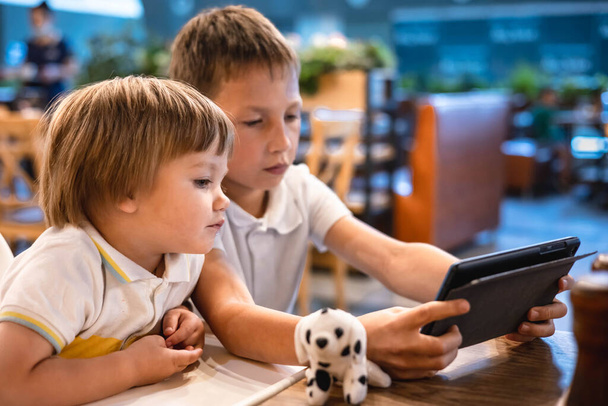 Çocuk ve bebek, restoranda siparişleri beklerken internetten tablette video oyunu oynuyorlar. Akıllı telefon kullanan çocuklar. Çocuklar teknolojiyi kullanıyor. Ekran zaman bağımlılığı. - Fotoğraf, Görsel