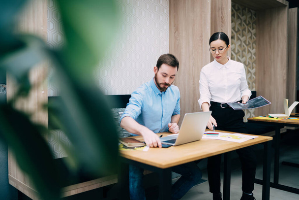 Young dark haired female in white blouse with printed paper document standing near male colleague in casual clothes sitting at workplace with laptop - Foto, immagini