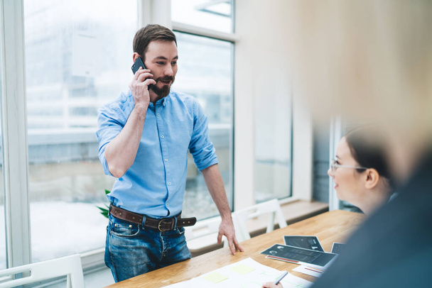 Positive bearded male in blue shirt jeans concentrating on speaking on smartphone standing opposite smiling female sitting at table in office - Фото, зображення