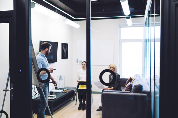 Widok przez szklane drzwi kobiet w formalnym zużyciu słuchanie wyjaśnień męskiego trenera biznesu przy użyciu białej planszy w biurze kreatywnym - Zdjęcie, obraz