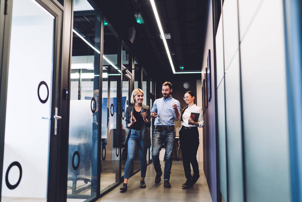 Young blonde woman in jacket and jeans and bearded happy man and dark haired female in glasses with notebooks smiling and walking through office corridor - Zdjęcie, obraz
