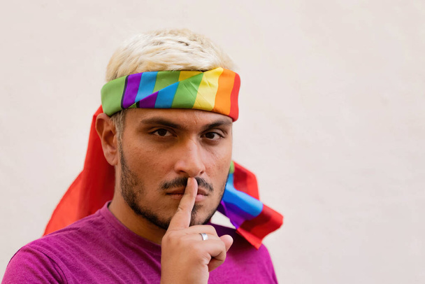 Bärtiger Typ mit LGBT-Fahne um den Kopf geschlungen, schaut in die Kamera und hält den Finger an den Lippen, während er vor beigem Hintergrund zum Schweigen auffordert - Foto, Bild