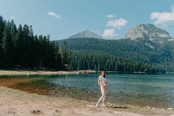 The girl looks at the Black Lake, forest and mountains in the Durmitor National Park of Montenegro. A tourist enjoys a magical view of the lake, a coniferous forest and a magical view, sitting on a - Foto, imagen