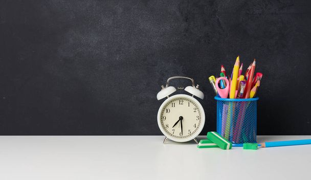 reloj despertador redondo blanco y un vaso de metal con bolígrafos, lápices y rotuladores en el fondo de una pizarra de tiza negra vacía, espacio para copiar - Foto, Imagen