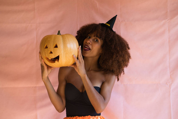 mladá africká Američanka oblečená jako čarodějka na halloween party a držící v rukou dýni. Nosí čarodějnický klobouk a oranžovou sukni. Žena se usmívá do kamery s dýní. - Fotografie, Obrázek