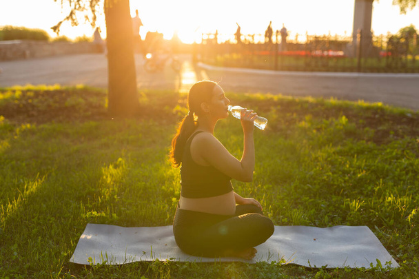 Femme enceinte boire de l'eau tout en étant assis sur un tapis de yoga dans le parc d'été. Mode de vie sain, concept de grossesse et de maternité. - Photo, image
