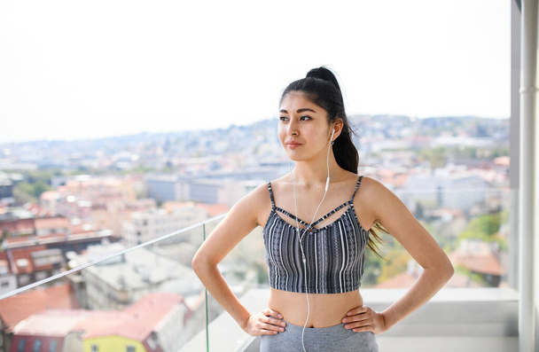 Portrait of young sport woman with earphones standing on balcony outdoors in city, resting. - Фото, изображение