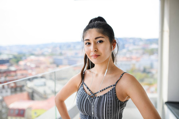 Portrait of young woman with earphones standing on balcony outdoors in city, looking at camera. - 写真・画像