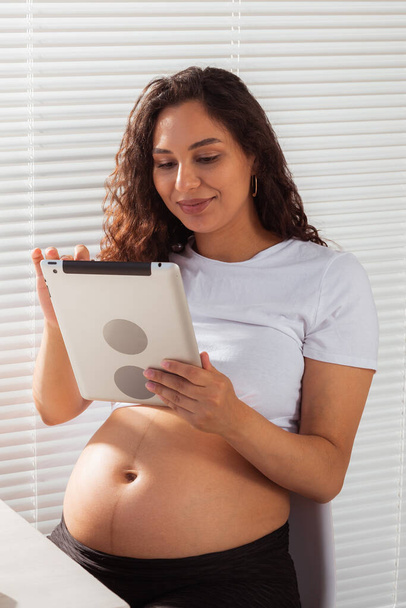 Hispanic pregnant woman using digital tablet while breakfast. Technology, pregnancy and maternity leave - Фото, зображення