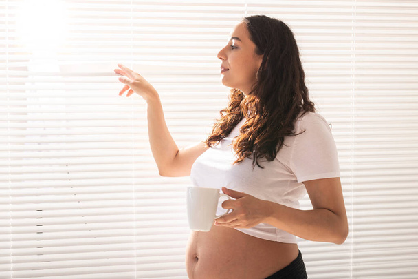 Zvědavá těhotná žena pije čaj a dívá se skrz žaluzie na okno. Koncept radosti a dobré zprávy při čekání na dítě - Fotografie, Obrázek