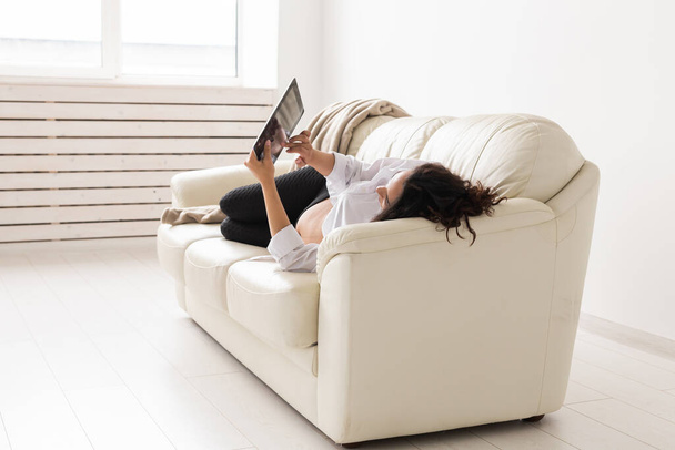 Spanisch schwangere Frau mit Tablet-Computer, während sie zu Hause auf dem Sofa liegt. Schwangerschaft und Informationen zum Elternschaftskonzept. - Foto, Bild