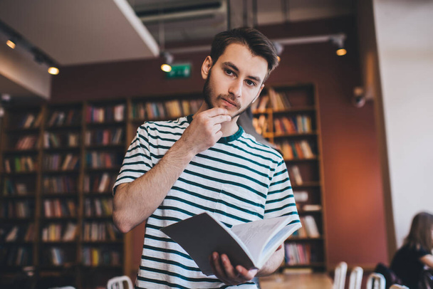 Pensive bearded male in casual outfit rubbing chin and thinking over book while standing in cozy library and looking at camera - Zdjęcie, obraz
