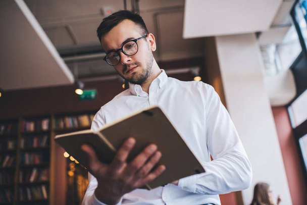 Low angle of male in white shirt and spectacles reading book and frowning while standing on blurred background of cozy bookshop - Photo, Image