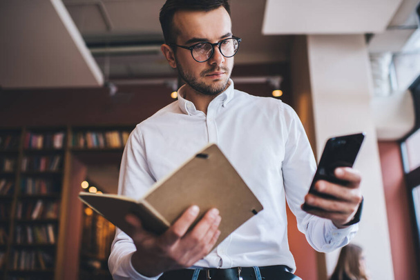 Low angle of serious male in glasses holding open book and browsing smartphone while standing on blurred background of library - Fotoğraf, Görsel