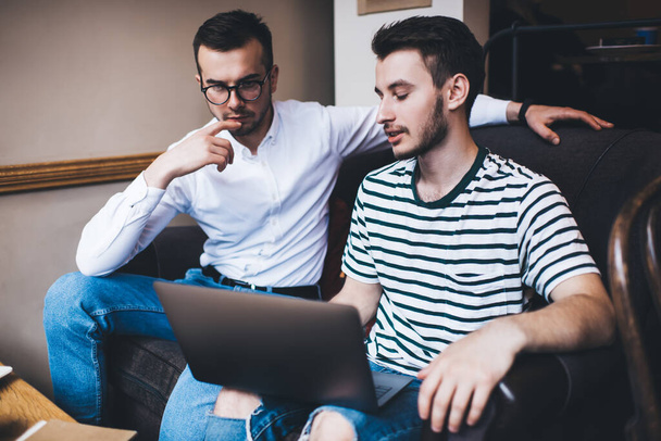Concentrated men in casual clothes having discussion and working on laptop while sitting on sofa by table with clipboard and notes - Foto, imagen