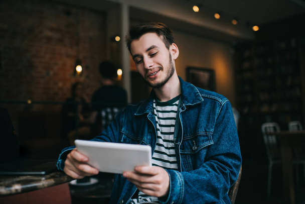 Young handsome freelance man in casual outfit sitting at table with laptop and smiling while browsing tablet in modern cafe  - 写真・画像