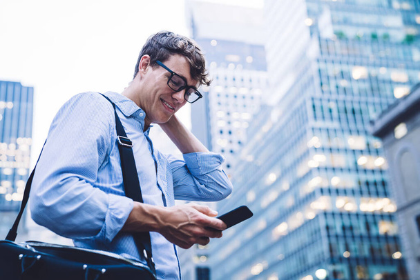 Low angle side view of man in shirt and eyeglasses messaging on phone standing against high buildings of New York looking surprised touching head  - 写真・画像