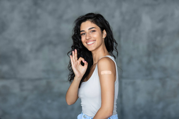 Ковидная вакцинация. Молодая женщина с лейкопластырем после инъекции коронавирусной вакцины жестом хорошо на сером фоне - Фото, изображение