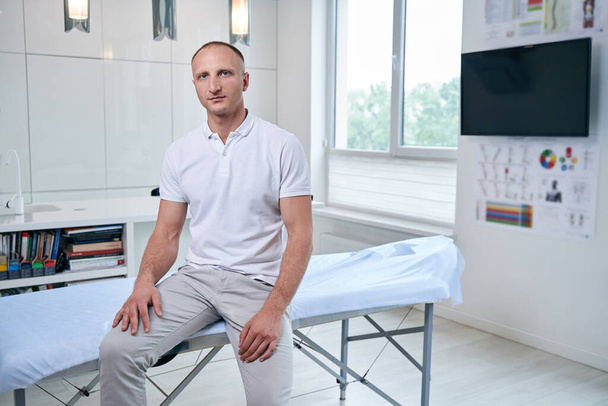Handsome Caucasian chiropractor sitting on massage table in medical office - Φωτογραφία, εικόνα
