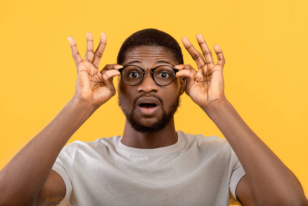 Emotionele zwarte man aanraken van zijn bril, schreeuwen WOW op gele studio achtergrond, man overweldigd door grote verkoop - Foto, afbeelding