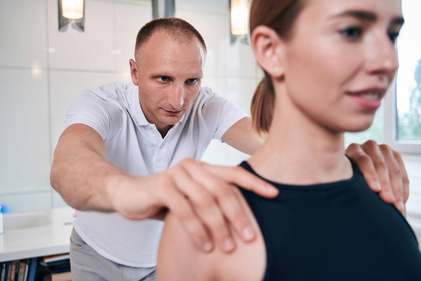 handsome neat physiotherapist examining shoulder of his woman client in medical office - Фото, зображення