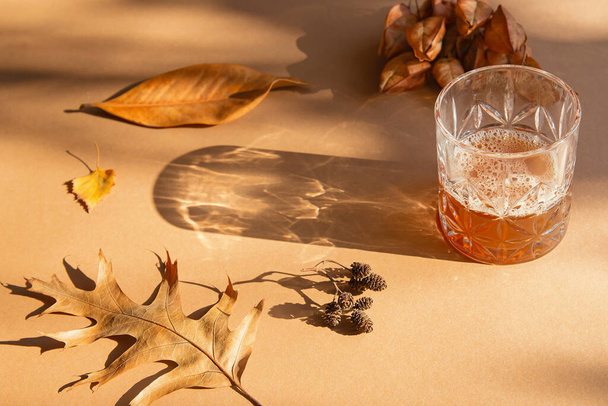 Glas met zelfgemaakte drank op lichtbruine zonovergoten achtergrond met gedroogde bladeren en schaduwen. Herfst- en herfstconcept - Foto, afbeelding