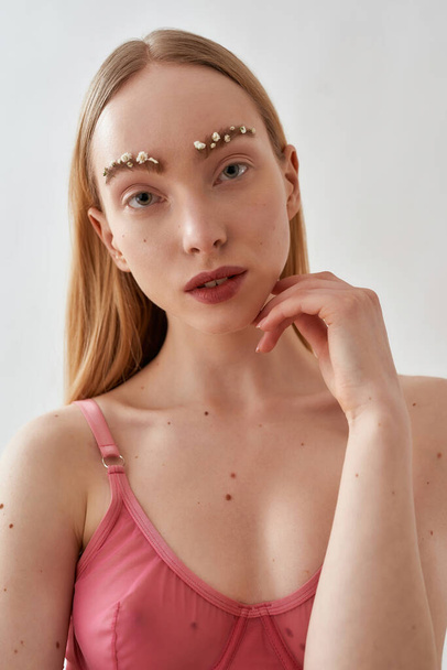 Closeup portrait of natural young caucasian woman with flowers in her eyebrows wearing pink transparent bra looking at camera, posing isolated over light gray background - Foto, imagen