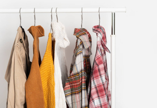 Autumn vintage clothing hangs on hangers on the rack. Beige trench coat, sweaters, plaid shirts. Organization of space, winter cleaning. - Photo, Image