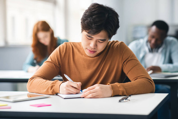 Студент-азиат сидит за столом в классе и пишет в блокнот - Фото, изображение