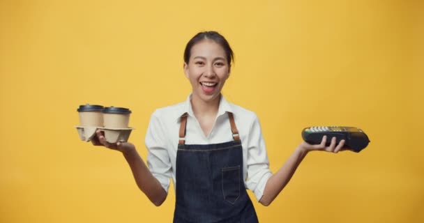 Szczęśliwy uśmiechnięty kelnerka Azji w fartuchu przyjazny patrząc na aparat dając kawę lub herbatę i czytnik kart kredytowych do klienta izolowane na żółtym tle. Koncepcja kawiarni dla przedsiębiorców. - Materiał filmowy, wideo