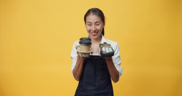 Gelukkig glimlachend Azië serveerster dragen schort vriendelijk op zoek naar camera geven koffie of thee en credit card reader machine aan klant geïsoleerd op de gele achtergrond. Entrepreneur cafe concept. - Video