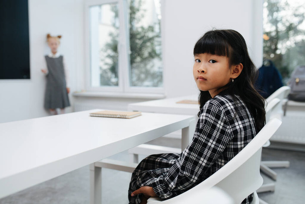Portrait of an asian schoolgirl in checkered uniform sitting behind the desk in a class. Looking at the camera. Blurred background. - Foto, Imagen