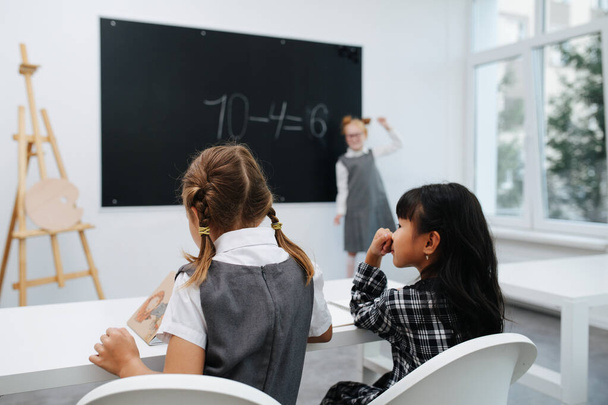 Two little schoolgirls sitting behind a desk, looking in a coorful book. Their classmate solving math equasions on a blackboard in a blurred background. - Photo, image