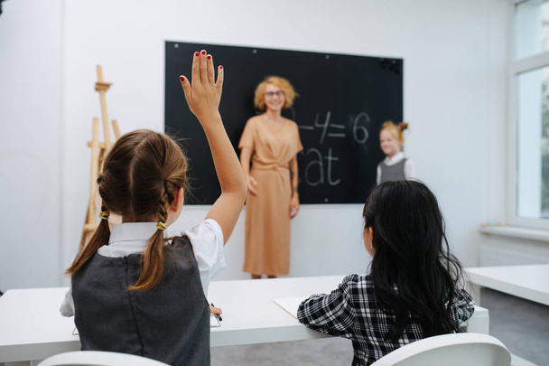 机の後ろに座っている2人の女の子、 1つに答えるために手を上げている。背景がぼやけている黒板の横に立っている. - 写真・画像