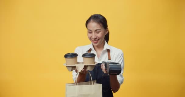 Szczęśliwy uśmiechnięty kelnerka Azji w fartuchu przyjazny patrząc na aparat dając kawę lub herbatę i czytnik kart kredytowych do klienta izolowane na żółtym tle. Koncepcja przedsiębiorstwa na wynos. - Materiał filmowy, wideo