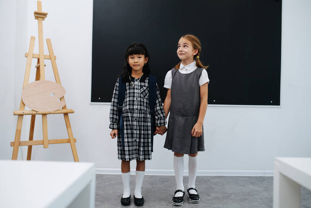 Two girl friends holding hands, standing in a class in front of a blackboard. Wearing different school uniform. - Foto, imagen