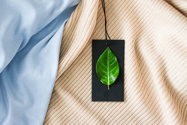 Зеленый лист на одежной бирке и органическом фоне ткани, устойчивая концепция моды и бренда этикетки - Фото, изображение