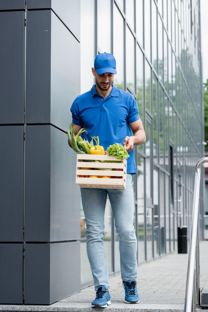 Uśmiechnięty arabski kurier z warzywami w pudełku spacerujący w pobliżu budynku na świeżym powietrzu - Zdjęcie, obraz