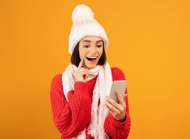Remote winter holiday shopping concept. Excited woman in knitted hat and scarf looking at smartphone, yellow background - Foto, imagen