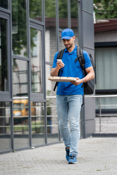Cheerful arabian deliveryman using smartphone while holding pizza box near building  - Photo, Image