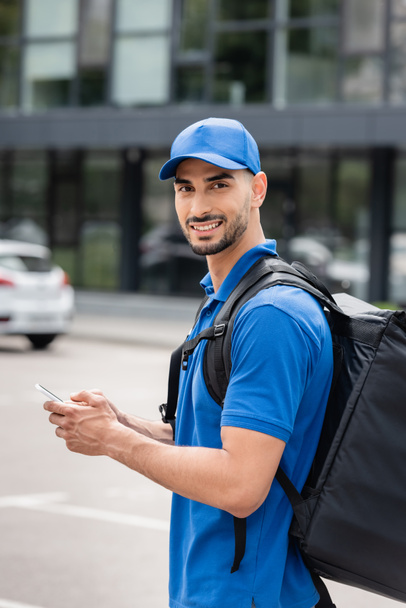 Young arabian courier with thermo backpack and smartphone looking at camera on urban street  - 写真・画像