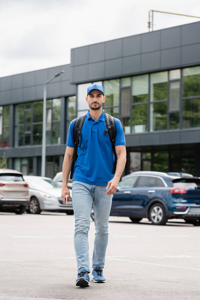 Arabian courier with backpack and smartphone walking on urban street  - Photo, Image