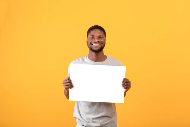 Fröhlicher afrikanisch-amerikanischer Mann mit weißem Blanko-Plakat, posiert und lächelt vor gelbem Studiohintergrund in die Kamera - Foto, Bild