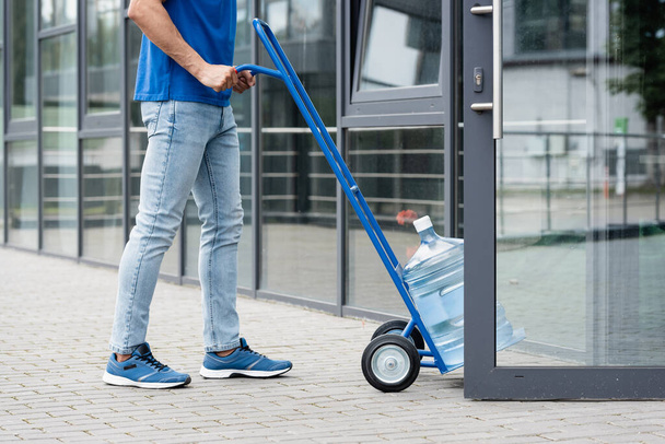 Cropped view of courier pulling cart with bottle of water near open door of building  - Fotoğraf, Görsel