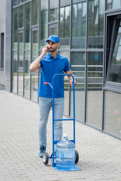 Arabian deliveryman in uniform talking on smartphone near bottle of water on cart and building  - 写真・画像