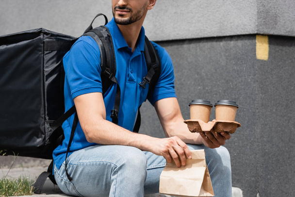 Cropped view of courier with thermo backpack holding paper bag and takeaway cups outdoors  - Fotografie, Obrázek