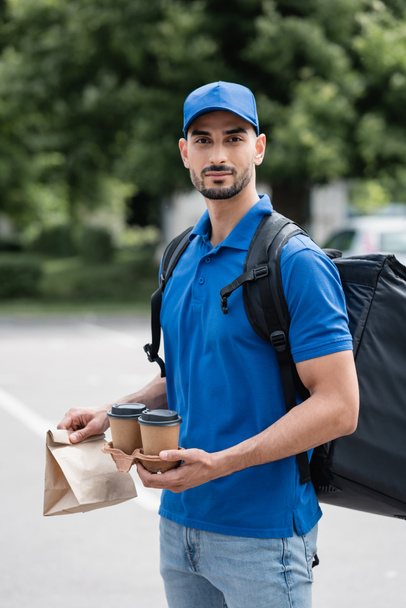 Termo sırt çantalı genç Mislim kurye elinde kahve, dışarıda kağıt torba bekliyor.  - Fotoğraf, Görsel